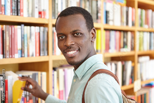 Indoor portrait of handsome African man picking book from shelve in bookshop as present on his friend's birthday. Black happy student spending break at college library, borrowing textbook for research