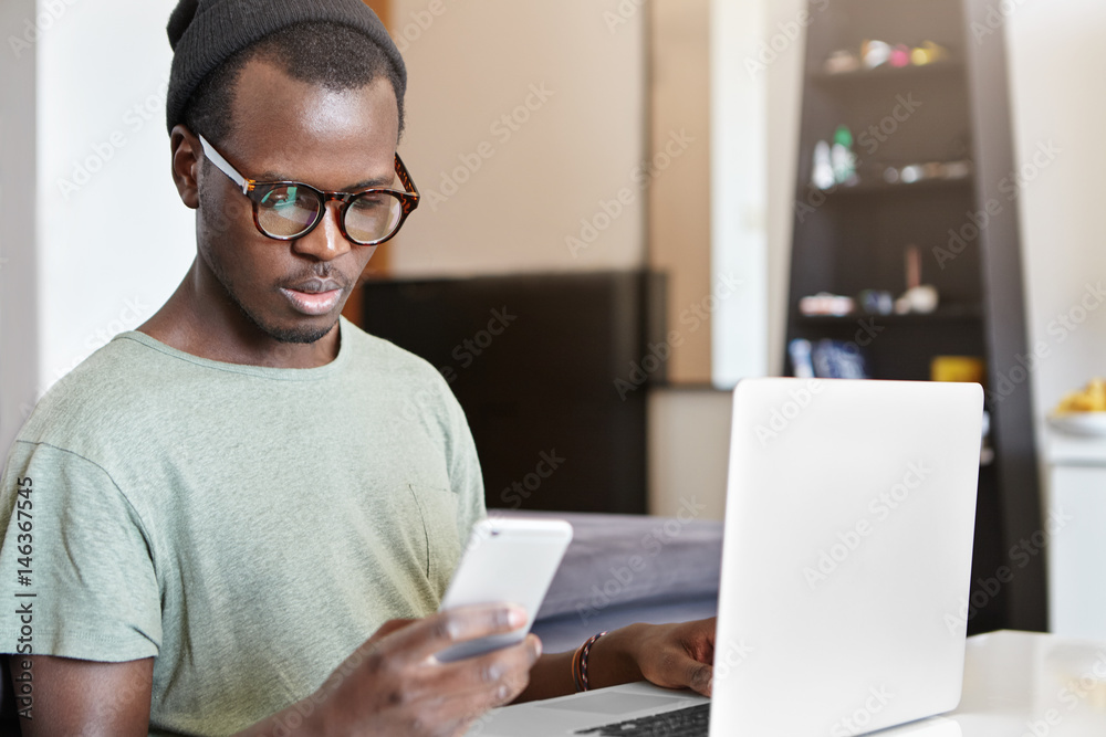 Staying connected. Stylish dark-skinned man using his smart phone for online banking, paying for wireless home internet while working on laptop computer. People, modern technology and communication Staying connected. Stylish dark-skinned man using his smart phone for online banking, paying for wireless home internet while working on laptop computer. People, modern technology and communication
