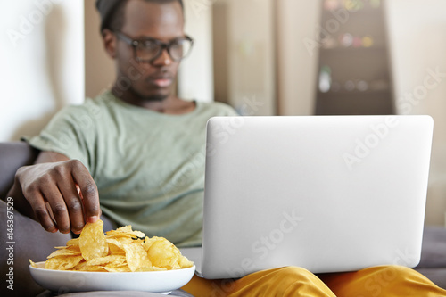 Candid shot of serious concentrated single male in hat and glasses relaxing in his apartment with laptop computer and plate of chips, surfing net or watching series. Selective focus on man's hand