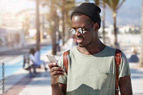 Stylish young Afro American male looking at screen of his smart phone with happy smile, about to answer his girlfriend's call while on vacations abroad. People, lifestye and modern communication photo