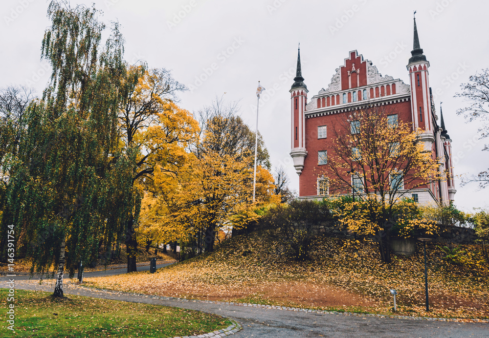 Obraz premium Admiralty House, golden trees and fallen leaves on Skeppsholmen islet in Stockholm by autumn cloudy day. Amiralitetshuset building near Skeppsholm bridge.