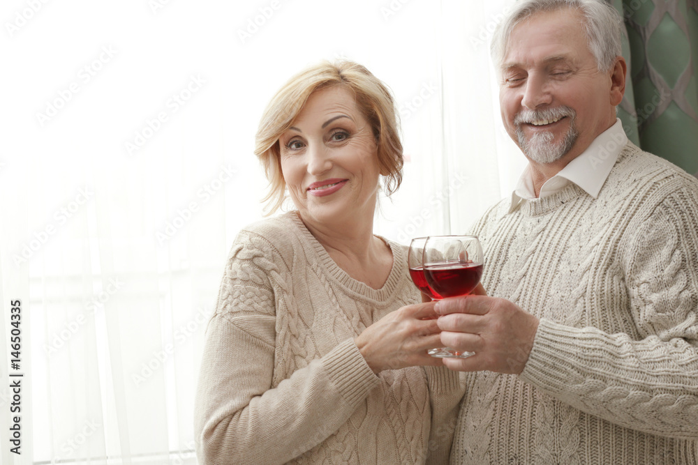 Cute elderly couple drinking wine while standing near window at home Cute elderly couple drinking wine while standing near window at home