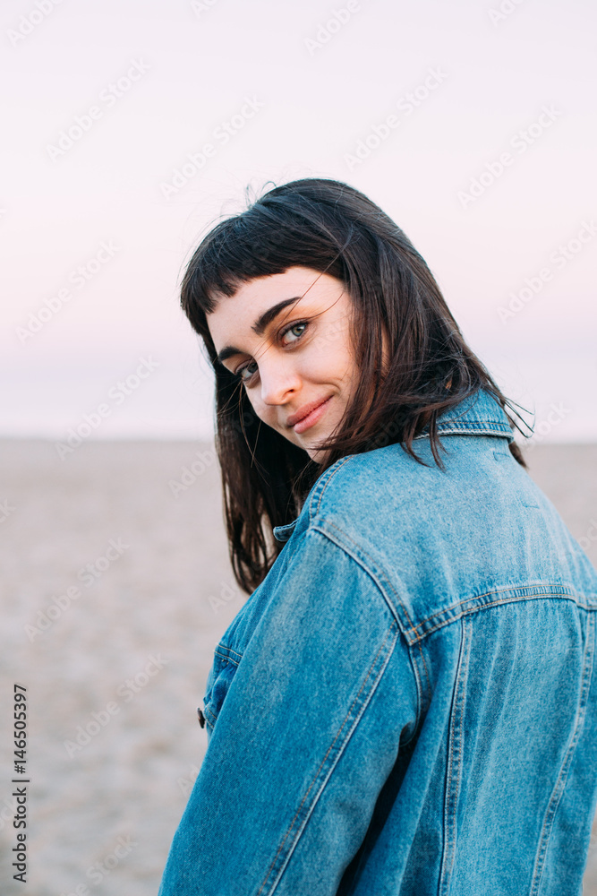 Fototapeta premium Cute dark haired russian girl poses during photoshoots on the beach with a pink sunset sky in the sand near the sea with the beach all smile filming and photograph portraits.