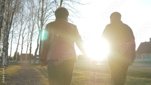 Loving elderly couple walking in the park at sunset. Nearby there is a road. A woman in a brown leather jacket. The man is dressed in a black sweater.