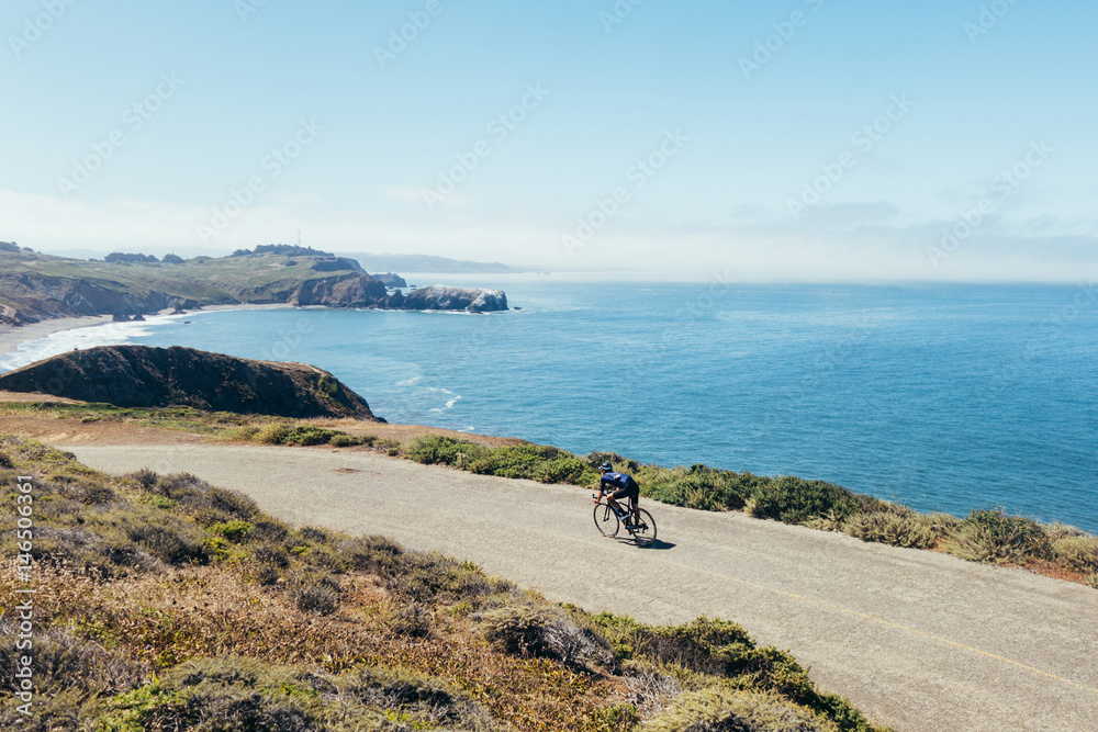 Obraz premium Slim fit strong male cyclist low aero riding fast on narrow road along the water toward beach in marin county, california.