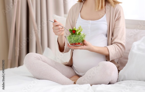 Young pregnant woman having breakfast while sitting on bed at home