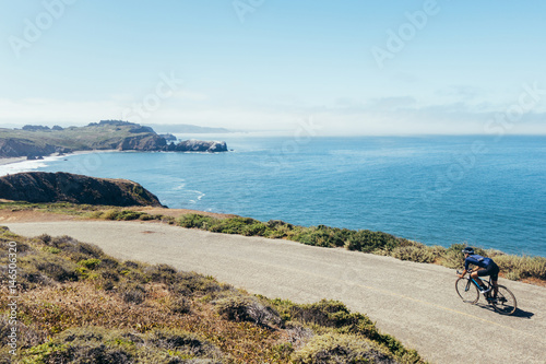 Slim fit strong male cyclist low aero riding fast on narrow road along the water toward beach in marin county, california.