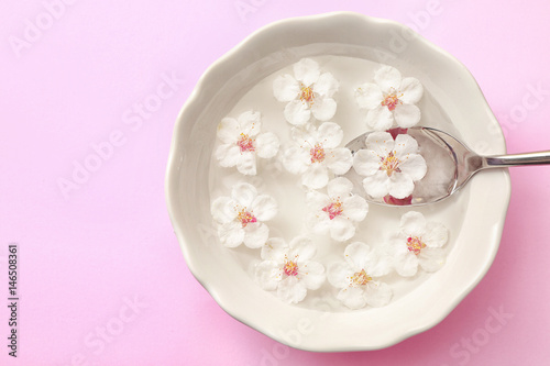 Bowl with fruit tree flowers, water and spoon on pink background
