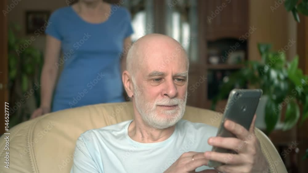 Senior couple at home. Husband using smartphone. Wife spying on her husband from his shoulder Senior couple at home. Husband using smartphone. Wife spying on her husband from his shoulder