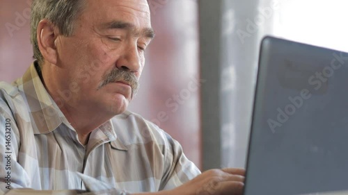 An elderly man with a mustache sits behind a laptop and solves problems. He looks seriously at the monitor