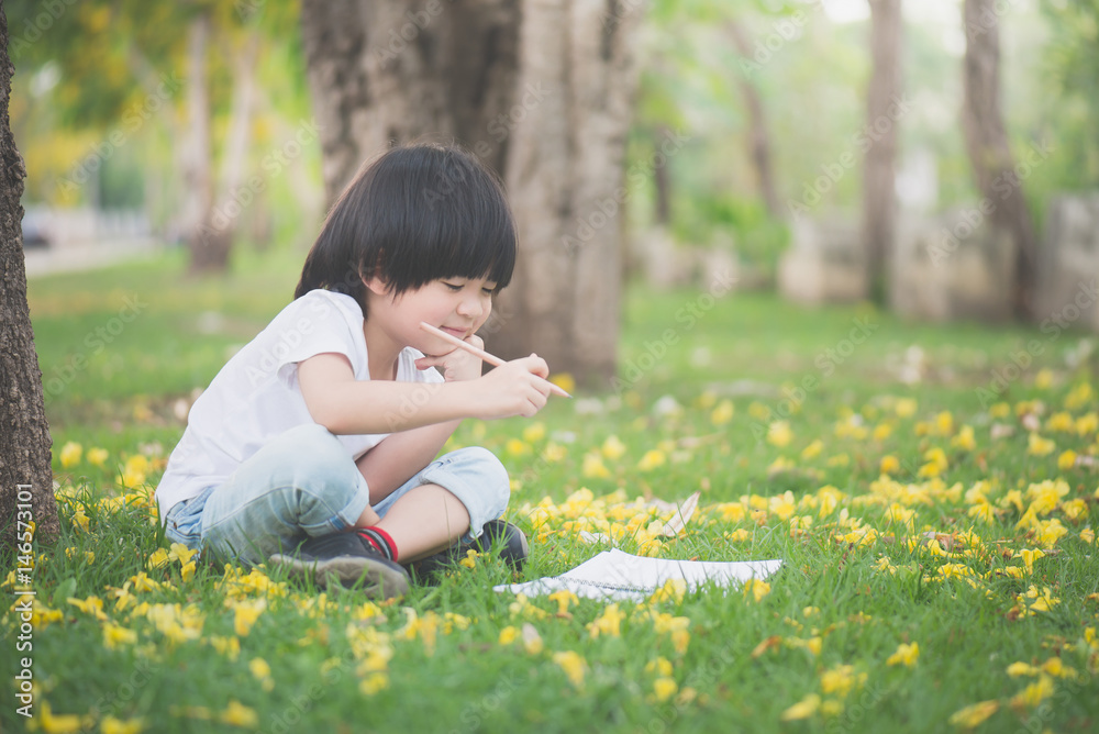 Little Asian boy sitting under the tree and drawing in notebook at park Little Asian boy sitting under the tree and drawing in notebook at park