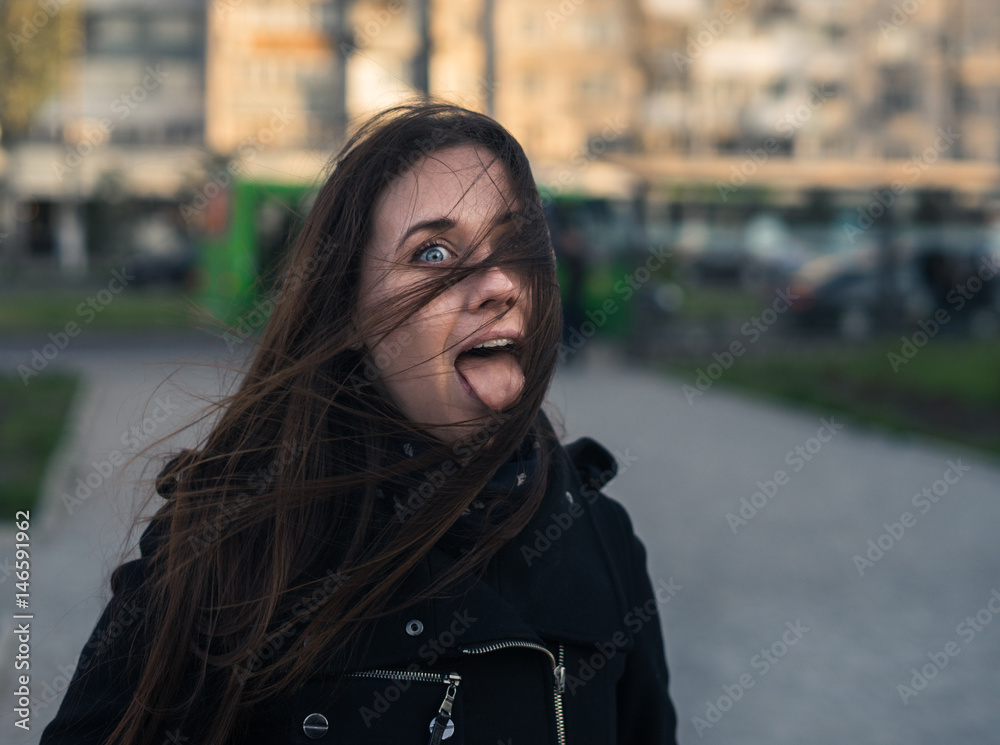 Crazy emotions of beautiful young girl in the city - she shows tongue. Strong wind blows her hair