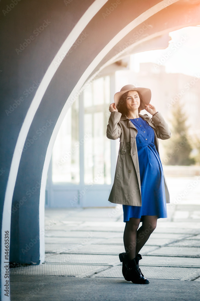 Fashionable pregnant woman with hat, classic coat, blue dress posing against a background of modern urban architecture