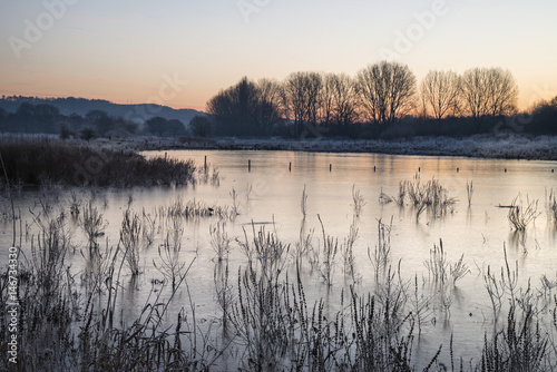 Beautiful vibrant English countryside lake image with frost and frozen lake in Winter at sunrise