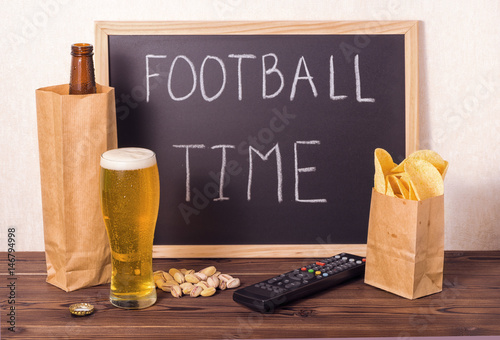 football fans setting of beer bottle in brown paper bag,  glass, chips, pistachio, tv remote control and handwriting text football time written in chalkboard over wooden table