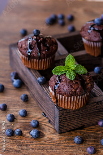 Chocolate muffins with chocolate syrup, blueberries and mint in a wooden background