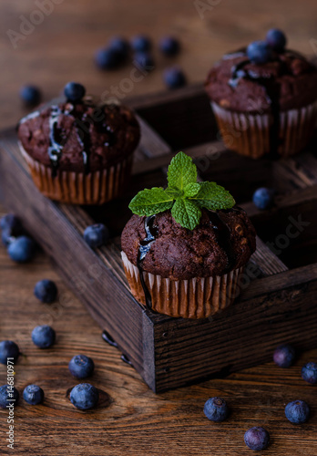 Chocolate muffins with chocolate syrup, blueberries and mint in a wooden background