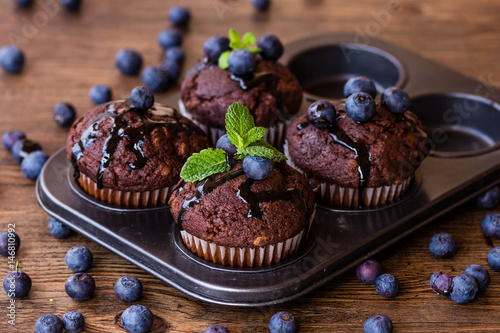 Chocolate muffins with chocolate syrup, blueberries and mint in a wooden background