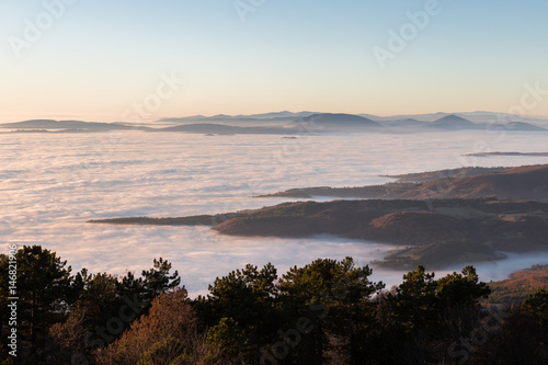 A sea of fog between some hills and some more distant mountains  with a line of trees in the foreground. It s sunset  so the colors are warm and soft