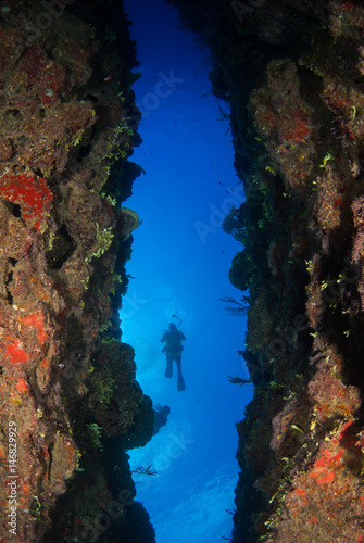 A diver swims through a large crack in the tropical reef which forms a sort of underwater passage. Coral can be seen in the foreground with the blue ocean above