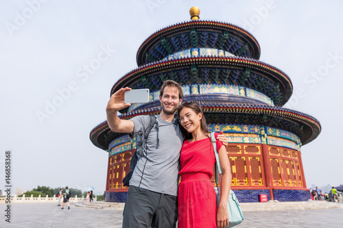 Happy couple travelers taking selfie picture together at temple of heaven during china summer travel. Young multiracial people using phone photography app for photos.