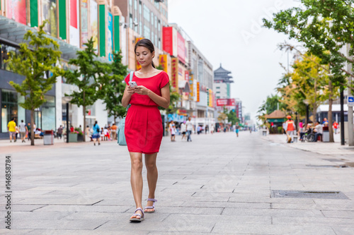 Asian woman walking downtown on Wangfujing shopping street in Beijing, china, using phone app to find directions or text online. Happy Chinese woman texting on mobile sms smartphone.