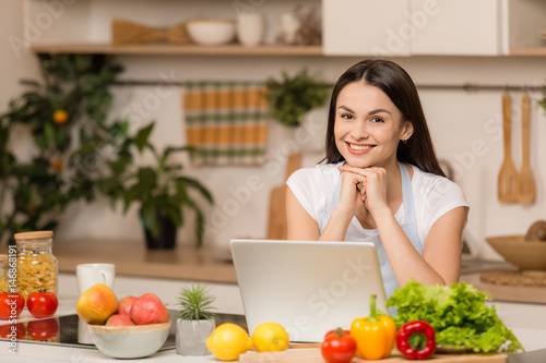 Young Woman in kitchen with laptop looking recipes, smiling. Food blogger concept