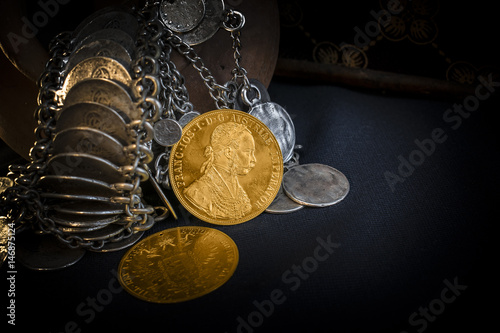Still Life with two Austria-Hungary thalers, avers and revers of golden coin-ducats from 1915 with Kaiser Franz Joseph I, leaning on silver jewelery and dark environment