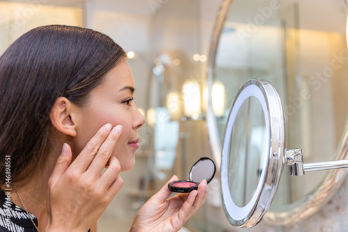 Canvas Print Asian young woman putting blush cream using fingers while looking in makeup mirror at home in bathroom