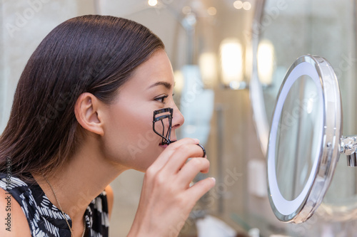 Woman using eyelash curler curling eyelashes before mascara in lighted round makeup mirror from luxury hotel or home bathroom. Beautiful Asian girl getting ready in the morning looking at reflection.
