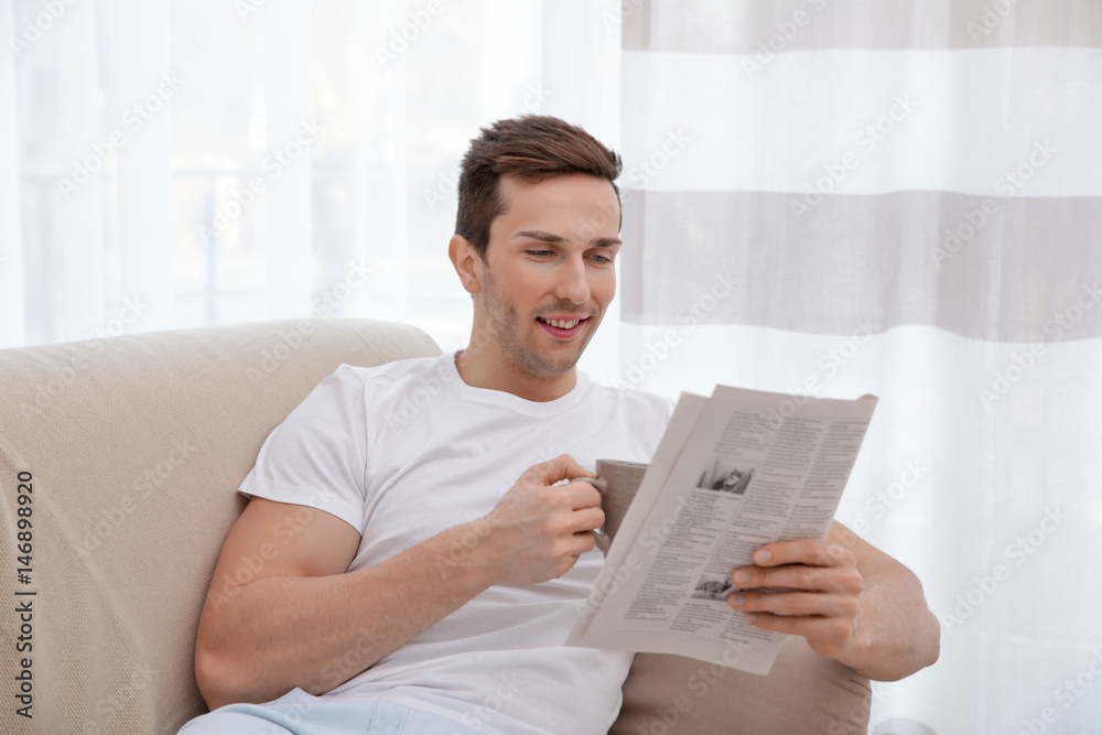 Young man resting on couch with newspaper and drinking tea in light room Young man resting on couch with newspaper and drinking tea in light room