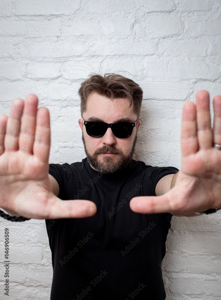 Naklejka premium Portrait of a bearded serious man wearing sunglasses and casual clothes on a white brick wall. He stretches his hands to the camera. The man performs the framing with his hands