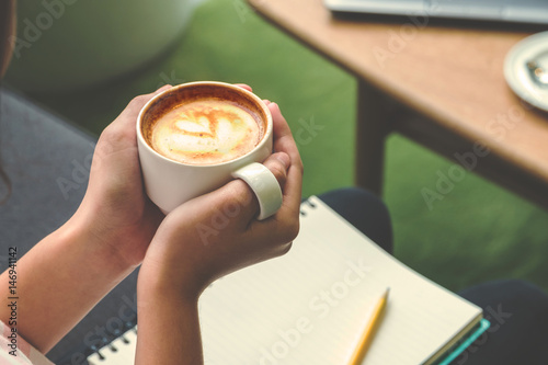 Close up hand holding hot cappuccino in white coffee cup with notebook and yellow pencil in cafe,Vintage filter