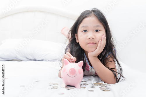 cute asian girl putting coin to piggy bank on bed.