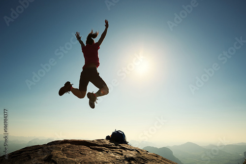 cheering successful young woman hiker jumping on mountain peak