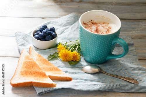 Breakfast idea. Cup with coffee with blueberries, dandelions and toast on linen napkin and wooden background