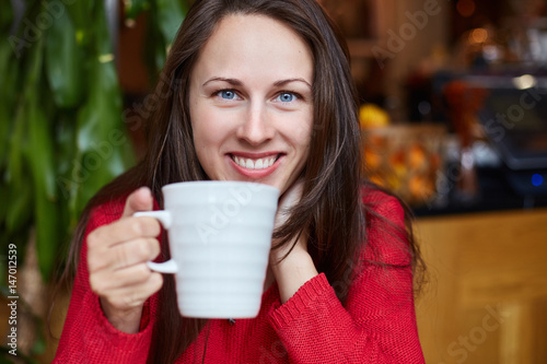 Tableau sur toile Young natural cheerful Caucasian brunette blue-eyed girl in red sweater is having her breakfast in modern organic coffee shop