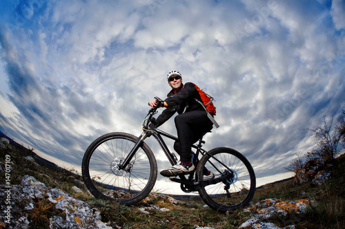 Portrait of the mountain cyclist standing with bike on the rocky hill against dramatic sky with clouds.