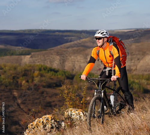 Young cyclist stands with the mountain bike on the summer rocky trail against beautiful landscape with mountain. © Aleksey