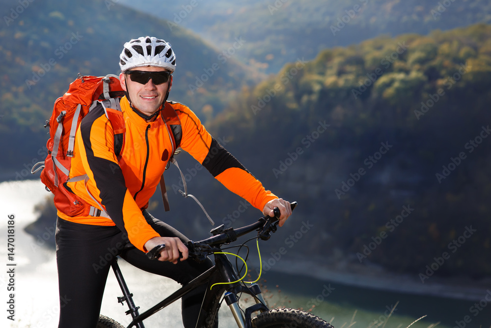 Portrait of young cyclist with a red backpack standing under river against summer landscape with mountain. Portrait of young cyclist with a red backpack standing under river against summer landscape with mountain.