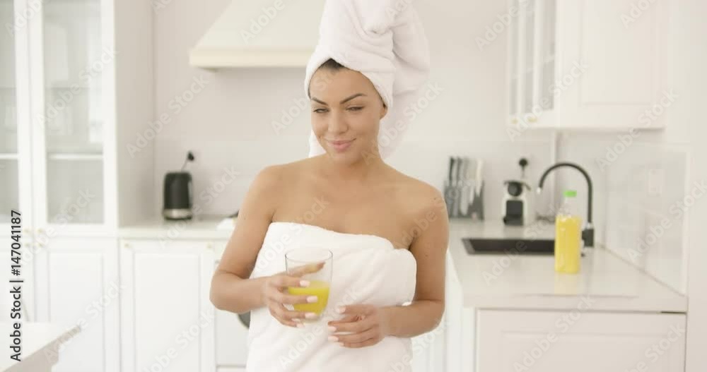 Young brunette wrapped in towel standing in light white kitchen holding glass of fresh orange juice and looking at camera smiling. Young brunette wrapped in towel standing in light white kitchen holding glass of fresh orange juice and looking at camera smiling.