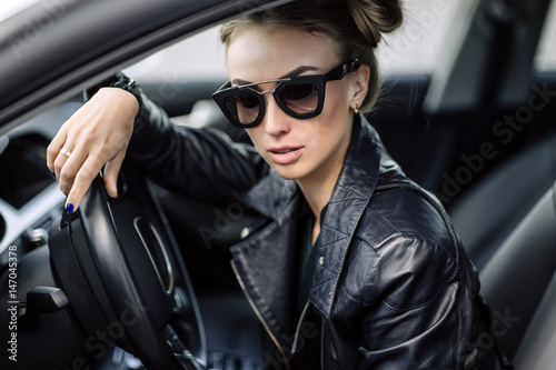 fashion outdoor photo of sexy beautiful woman with dark hair in black leather jacket and sunglasses posing in luxurious auto © boykovi1991