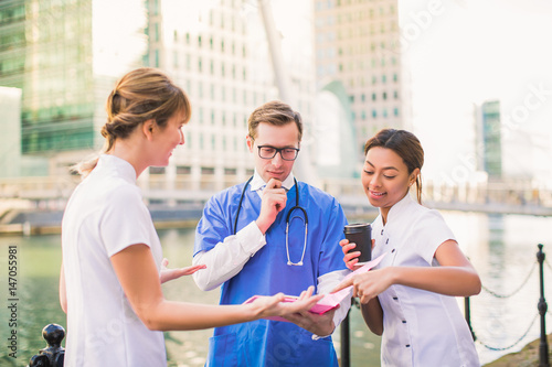 Three doctors stand and look at folder, discuss about something, in time of break, outdoors