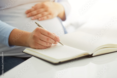 Young pregnant woman writes to her diary on a white table
