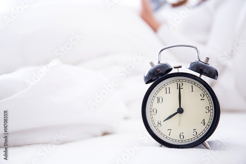 alarm clock standing on bedside table going to ring early morning to wake up woman in bed sleeping in background - early awakening -not getting enough sleep - getting work time concept