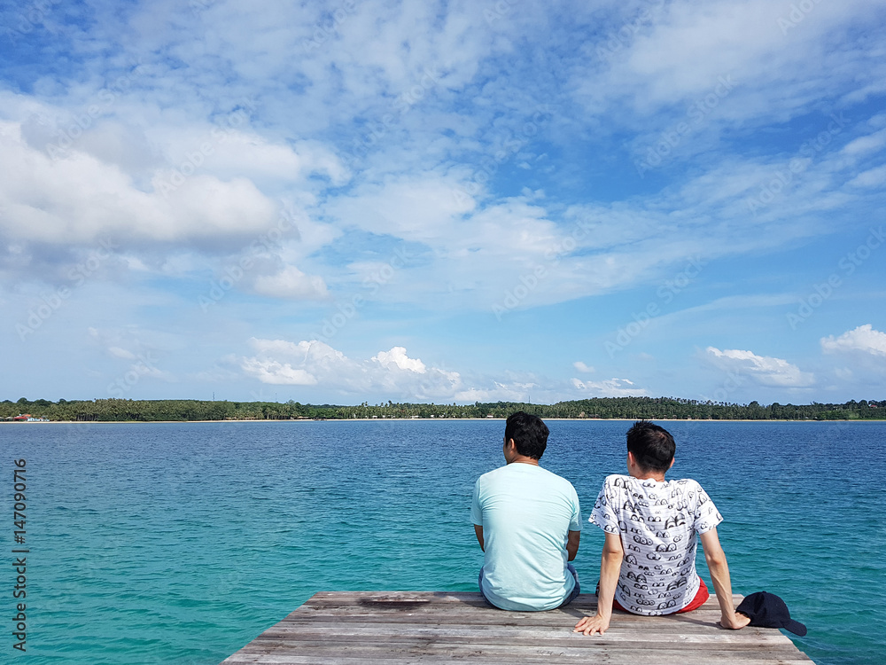 Two man sitting together on pier of wooden bridge in the sea and looking forward