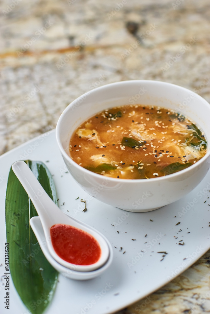 Korean cuisine. Spicy kimchi soup in white ceramic bowl served with red sauce in spoon, green leaf and paprika rings with glass of white wine on marble table. Korean cuisine. Spicy kimchi soup in white ceramic bowl served with red sauce in spoon, green leaf and paprika rings with glass of white wine on marble table.