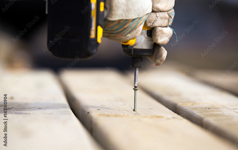 Close up of hands wearing work gloves gripping a power drill putting a screw into a piece of wood Close up of hands wearing work gloves gripping a power drill putting a screw into a piece of wood