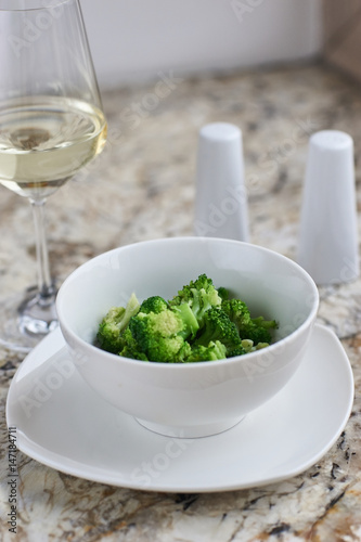 Steamed broccoli served in white bowl with glass of white wine on marble background