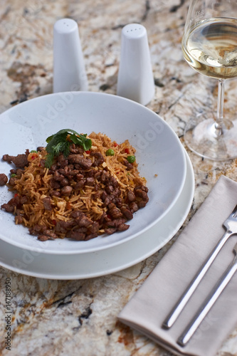 Fried rice with beef slices served in white round plate with glass of white wine on marble table